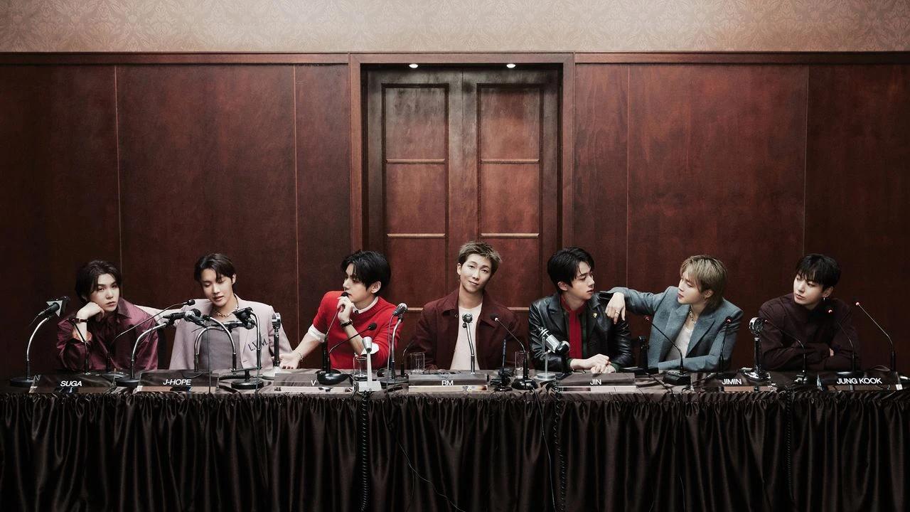 All seven BTS members seated at a press conference table with microphones, photographed by Dukhwa Jang for GQ March 2026